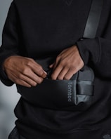 A person wearing a black sweatshirt is seen with their hands inside a small black bag with a shoulder strap. The bag has a logo printed on it.