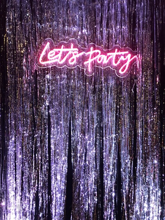A vibrant neon sign that reads 'Let's Party' illuminated against a backdrop of shiny tinsel or metallic streamers. The neon sign emits a bright pink glow that contrasts with the purple and silver hues of the streamers, creating a festive and celebratory atmosphere.