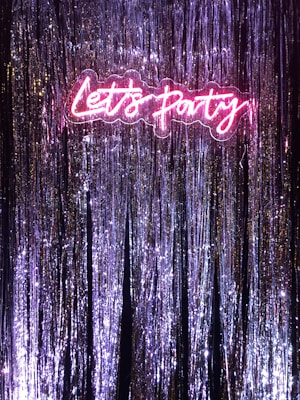 A vibrant neon sign that reads 'Let's Party' illuminated against a backdrop of shiny tinsel or metallic streamers. The neon sign emits a bright pink glow that contrasts with the purple and silver hues of the streamers, creating a festive and celebratory atmosphere.