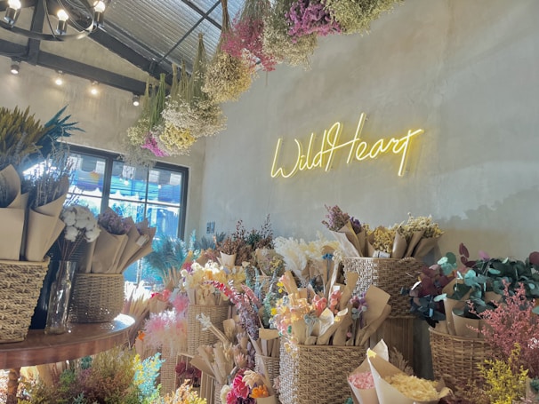 A cozy floral shop interior with bouquets of dried flowers arranged in baskets and wrapped in brown paper. A neon sign on the wall spells 'Wild Heart' in glowing yellow. The ceiling has hanging dried flowers, and natural light filters through a window, creating a warm and inviting atmosphere.