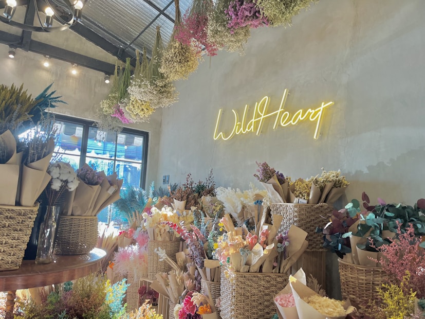 A cozy floral shop interior with bouquets of dried flowers arranged in baskets and wrapped in brown paper. A neon sign on the wall spells 'Wild Heart' in glowing yellow. The ceiling has hanging dried flowers, and natural light filters through a window, creating a warm and inviting atmosphere.
