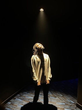 A person stands under a spotlight in a dark room, looking upward with a thoughtful expression. The light casts a warm glow on the individual's face and upper body, while the surroundings remain dimly lit.