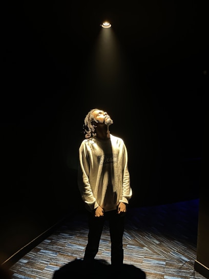 A person stands under a spotlight in a dark room, looking upward with a thoughtful expression. The light casts a warm glow on the individual's face and upper body, while the surroundings remain dimly lit.