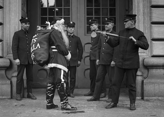 A group of men in uniform stand in front of a large building entrance, surrounding a person dressed as Santa Claus carrying a large sack. One man holds a fire hose nozzle as if aiming it towards Santa.