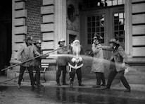 A group of firefighters dressed in traditional gear stand around a person in a Santa Claus outfit. They appear to be playfully spraying water from a hose towards Santa in front of a brick fire station building. The scene has a jovial and playful air, capturing a moment of lighthearted interaction.