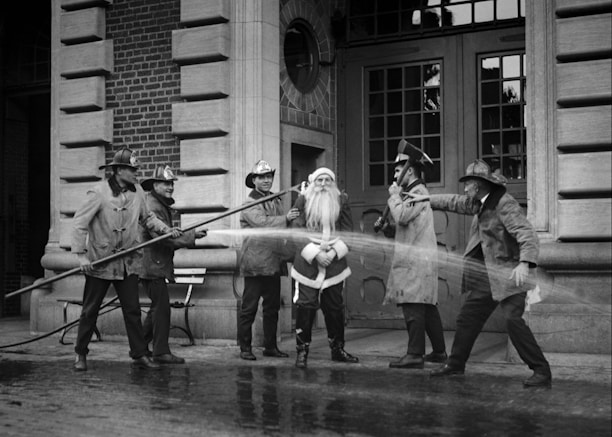 A group of firefighters dressed in traditional gear stand around a person in a Santa Claus outfit. They appear to be playfully spraying water from a hose towards Santa in front of a brick fire station building. The scene has a jovial and playful air, capturing a moment of lighthearted interaction.