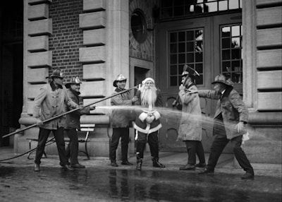 A group of firefighters dressed in traditional gear stand around a person in a Santa Claus outfit. They appear to be playfully spraying water from a hose towards Santa in front of a brick fire station building. The scene has a jovial and playful air, capturing a moment of lighthearted interaction.