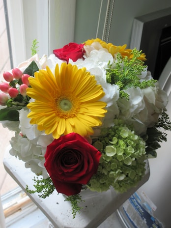A vibrant bouquet featuring a prominent yellow sunflower, surrounded by red roses, white hydrangeas, and small clusters of pink berries. Greenery adds contrast to the arrangement, which sits on a white surface near a window.