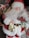 A Santa Claus figure dressed in traditional red and white attire with a fluffy white beard and a large gold buckle on its black belt. He is holding a gold-colored sack and a decorated gift. There is a festive arrangement with shiny ornaments and greenery in the background.