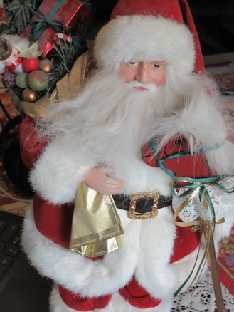 A Santa Claus figure dressed in traditional red and white attire with a fluffy white beard and a large gold buckle on its black belt. He is holding a gold-colored sack and a decorated gift. There is a festive arrangement with shiny ornaments and greenery in the background.