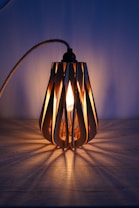 A decorative lamp with a wooden, slatted design emits a warm glow. The light bulb inside creates a pattern of shadows on the surrounding surface. The lamp is placed on a wooden surface, and the background is softly illuminated, adding to the cozy ambiance.