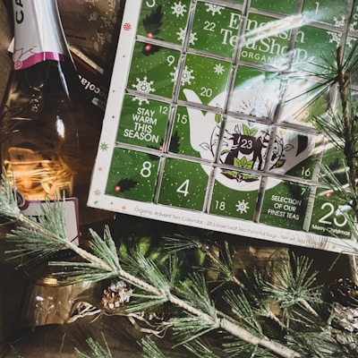 A collection of holiday-themed items, including a green and white advent calendar marked with numbers and festive motifs such as snowflakes. A bottle of sparkling wine is partially visible next to the calendar, accompanied by decorative evergreen branches and a small pine cone.