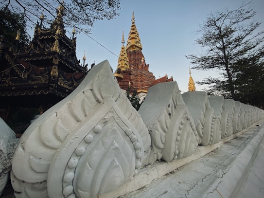 An ornate Buddhist temple features intricate architectural designs, with a striking gold spire rising prominently amidst a clear blue sky. In the foreground, there is a detailed stone balustrade with leaf-like patterns. Trees partially frame the structure, adding a touch of nature to the setting.