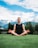 A person is meditating in a seated position on a grassy field. The background shows snow-capped mountains under a partly cloudy sky.