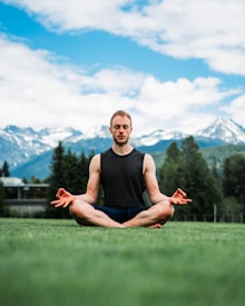 A person is meditating in a seated position on a grassy field. The background shows snow-capped mountains under a partly cloudy sky.