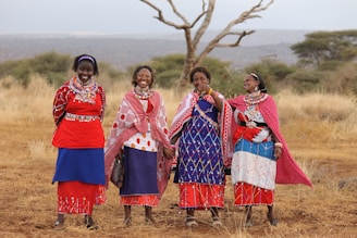 Four women stand together outdoors, wearing vibrant traditional attire adorned with colorful beads and patterns. They are smiling and appear to be enjoying a moment together in a natural setting. The landscape features dry grass and sparse trees under a vast, open sky.