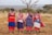 Four women stand together outdoors, wearing vibrant traditional attire adorned with colorful beads and patterns. They are smiling and appear to be enjoying a moment together in a natural setting. The landscape features dry grass and sparse trees under a vast, open sky.