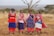 Four women stand together outdoors, wearing vibrant traditional attire adorned with colorful beads and patterns. They are smiling and appear to be enjoying a moment together in a natural setting. The landscape features dry grass and sparse trees under a vast, open sky.