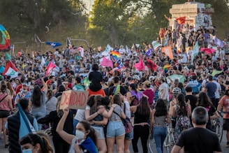 A large crowd of people gathered in an outdoor setting, holding various colorful flags and banners with words and symbols. Some individuals are sitting or standing on a monument or platform. The atmosphere is dynamic with many people seemingly engaged in a protest or public demonstration.