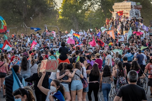 A large crowd of people gathered in an outdoor setting, holding various colorful flags and banners with words and symbols. Some individuals are sitting or standing on a monument or platform. The atmosphere is dynamic with many people seemingly engaged in a protest or public demonstration.