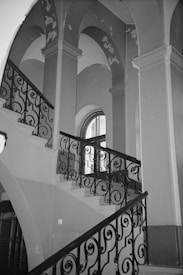 An ornate staircase with intricately designed wrought iron railings ascends within a building featuring tall, arched ceilings. The walls show signs of wear, with patches of peeling paint visible. A large, arched window is situated at the upper level, allowing some light into the space.