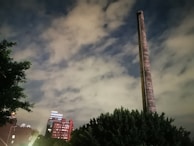 A tall, cylindrical chimney stands prominently against a cloudy sky, with residential or commercial buildings visible in the background. Trees frame the lower part of the image, adding a touch of greenery.