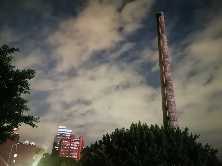 A tall, cylindrical chimney stands prominently against a cloudy sky, with residential or commercial buildings visible in the background. Trees frame the lower part of the image, adding a touch of greenery.