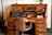An antique wooden roll-top desk featuring a vintage typewriter and early office equipment. The desk has multiple small drawers and compartments, with a large wooden chair positioned in front. Additional items on the desk include an old telephone, inkwells, and paper.