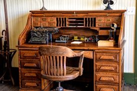 An antique wooden roll-top desk featuring a vintage typewriter and early office equipment. The desk has multiple small drawers and compartments, with a large wooden chair positioned in front. Additional items on the desk include an old telephone, inkwells, and paper.