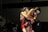 Two wrestlers are engaged in a competitive match. The athlete in red holds a strong grip on the opponent from behind. They both wear protective headgear, and their expressions indicate intense focus. The background shows a dimly lit gymnasium with a few spectators.