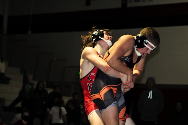 Two wrestlers are engaged in a competitive match. The athlete in red holds a strong grip on the opponent from behind. They both wear protective headgear, and their expressions indicate intense focus. The background shows a dimly lit gymnasium with a few spectators.