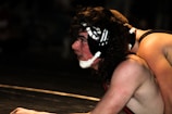 Two wrestlers are engaged in a match. One athlete is on top of the other, both wearing headgear and singlets. The wrestler on the bottom has long hair and appears focused and determined.