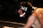 Two wrestlers are engaged in a match. One athlete is on top of the other, both wearing headgear and singlets. The wrestler on the bottom has long hair and appears focused and determined.