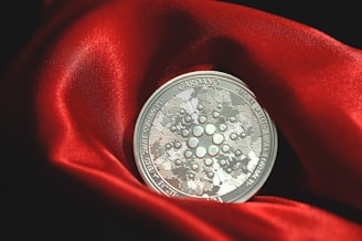 A silver Cardano coin rests on a rich, red satin fabric. The intricate design of the coin displays the name 'Cardano' and features circuit-like patterns, conveying a sense of technology and innovation. The fabric's texture adds a luxurious context and contrasts with the metallic sheen of the coin.