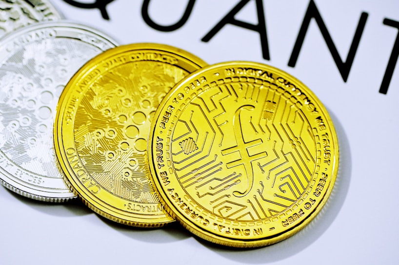 Three cryptocurrency coins are placed on a surface. The forefront coin is gold with a detailed circuit-like pattern and inscriptions. The coin in the background is silver, featuring similar intricate designs. A portion of a word starting with 'QUANT' is visible in the top background.
