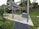 A wooden pergola structure located in a park setting. There is a concrete pathway with surrounding grass and small white flowers. Bushes and trees are in the background. A gray barbecue unit is positioned under the pergola.