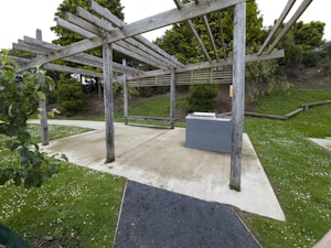 A wooden pergola structure located in a park setting. There is a concrete pathway with surrounding grass and small white flowers. Bushes and trees are in the background. A gray barbecue unit is positioned under the pergola.