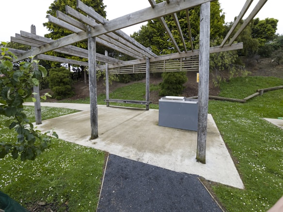 A wooden pergola structure located in a park setting. There is a concrete pathway with surrounding grass and small white flowers. Bushes and trees are in the background. A gray barbecue unit is positioned under the pergola.