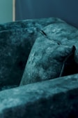 A close-up view of a plush, textured, dark teal sofa with a soft velvet-like fabric. The focus is on the well-cushioned corner, which appears inviting and comfortable.