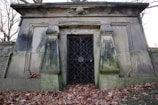 A stone mausoleum with a weathered appearance, featuring two prominent decorative columns and a metal gate. The structure is surrounded by bare trees and fallen autumn leaves scattered on the ground.
