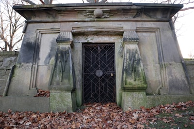 A stone mausoleum with a weathered appearance, featuring two prominent decorative columns and a metal gate. The structure is surrounded by bare trees and fallen autumn leaves scattered on the ground.
