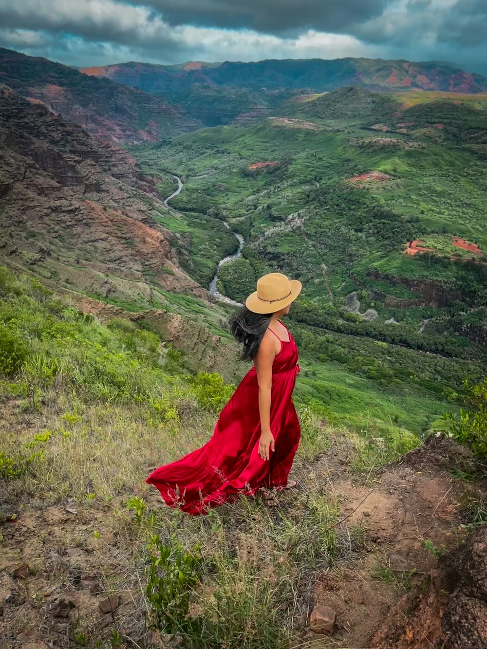 a woman in a red dress and a hat standing on a hill