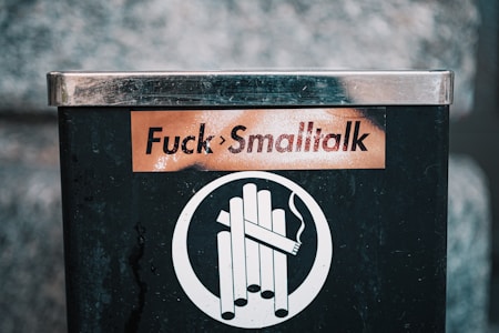 A black and silver cigarette disposal bin with a sticker that reads 'Fuck Smalltalk' in bold letters. Below the sticker, there is a symbol indicating cigarettes.