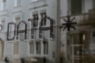 Text formed by small dots spelling the word 'DATA' followed by an asterisk, seen on a glass surface with a blurred building in the background.