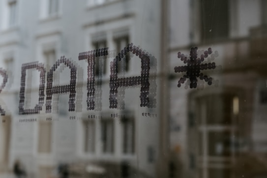 Text formed by small dots spelling the word 'DATA' followed by an asterisk, seen on a glass surface with a blurred building in the background.