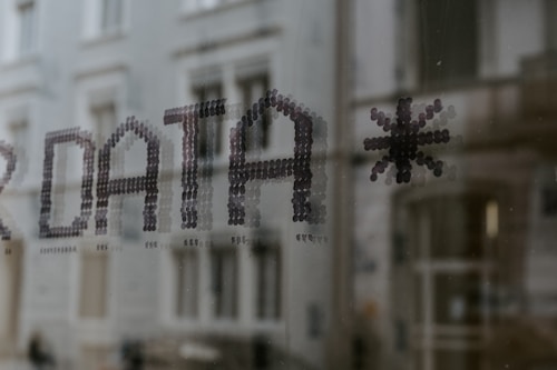 Text formed by small dots spelling the word 'DATA' followed by an asterisk, seen on a glass surface with a blurred building in the background.