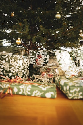 A collection of wrapped gifts sits beneath a decorated Christmas tree. The presents are adorned with green and white patterned wrapping paper and are tied with red ribbons. The tree is adorned with a few delicate ornaments.