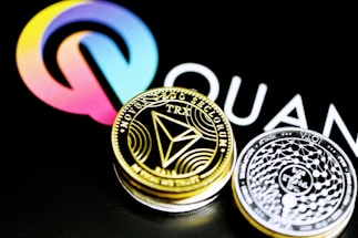 A close-up of two cryptocurrency coins, one gold-colored featuring the TRON (TRX) symbol and the other silver-colored with intricate designs, placed against a background displaying a colorful logo and partial text.