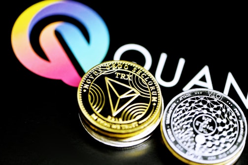 A close-up of two cryptocurrency coins, one gold-colored featuring the TRON (TRX) symbol and the other silver-colored with intricate designs, placed against a background displaying a colorful logo and partial text.