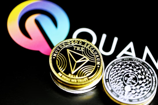 A close-up of two cryptocurrency coins, one gold-colored featuring the TRON (TRX) symbol and the other silver-colored with intricate designs, placed against a background displaying a colorful logo and partial text.